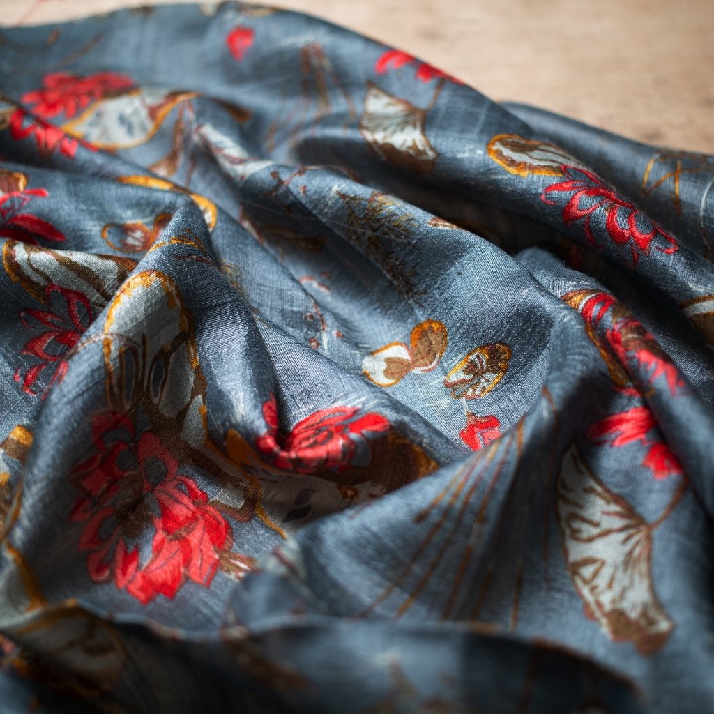A close-up shot of a blue printed organza fabric with colorful floral and butterfly patterns in red, orange, and white, showing light sheen and delicate folds.