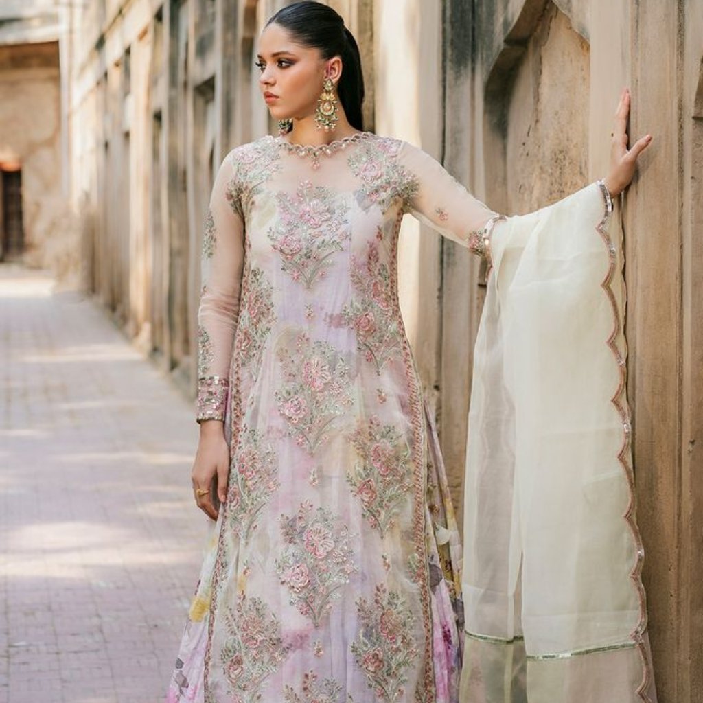 A stylish woman wearing a pastel embroidered organza Anarkali dress with floral embroidery, sheer sleeves, and a matching sheer dupatta, standing against a stone wall in a sunlit alley.