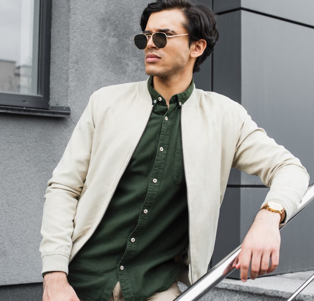 A fashion-forward man wearing a light beige bomber jacket over a dark green button-up shirt, sunglasses, and a gold watch. He leans on a metal railing outdoors against a modern gray building.