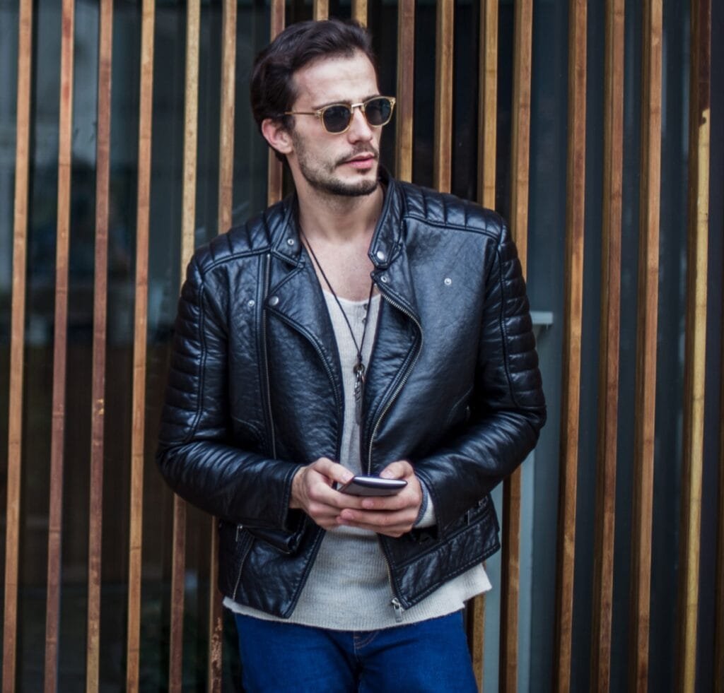 A stylish man wearing a black leather bomber jacket, sunglasses, and a light-gray knit shirt, standing against a wooden slat wall while looking off to the side and holding a smartphone.