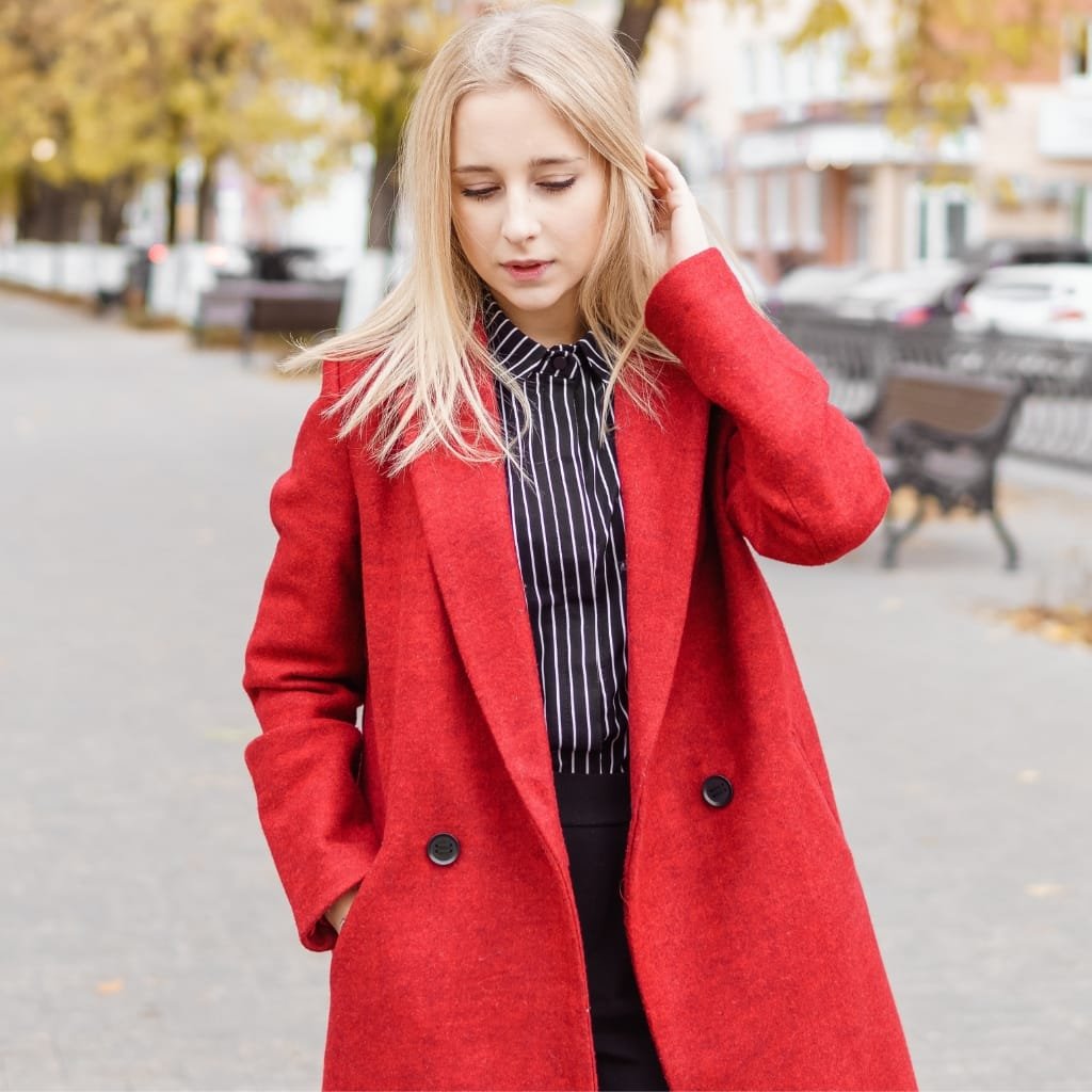 A young woman with blonde hair walks outdoors on a city street in autumn. She wears a bright red wool double-breasted coat over a black-and-white pinstripe shirt, with her hands in her coat pockets. The background shows a sidewalk, benches, and fall trees with yellow leaves.
