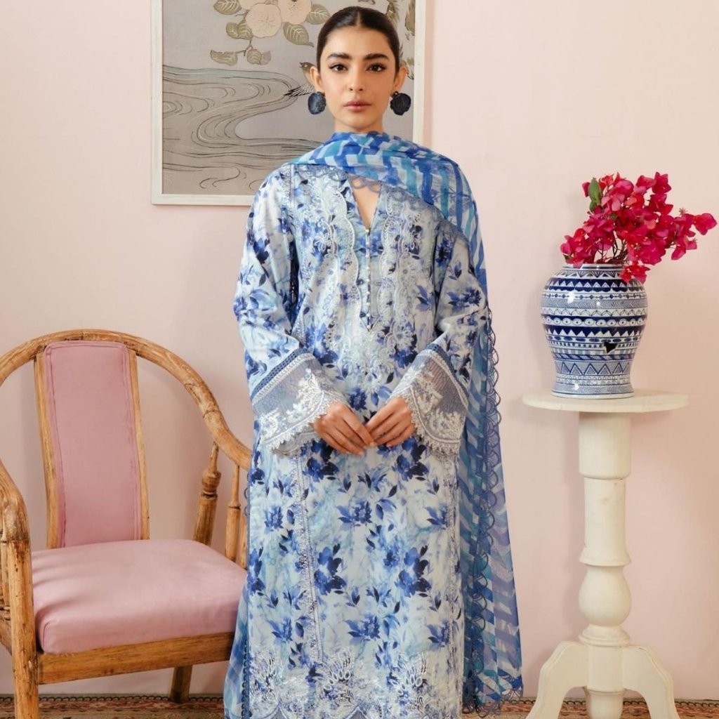 A woman stands indoors wearing a blue and white printed organza dress with lace details. She has a matching blue scarf draped over her shoulders. The room features a pink wall, a wooden chair with a pink cushion, a white pedestal table, and a blue-and-white ceramic vase with pink flowers.