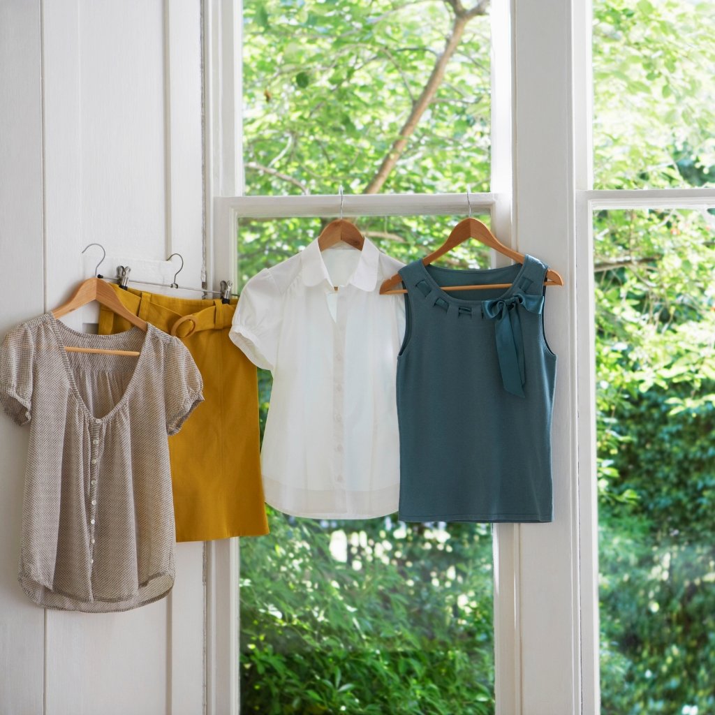 A clothesline of four garments hanging on wooden hangers in front of a bright window with greenery outside. lawn cotton fabric