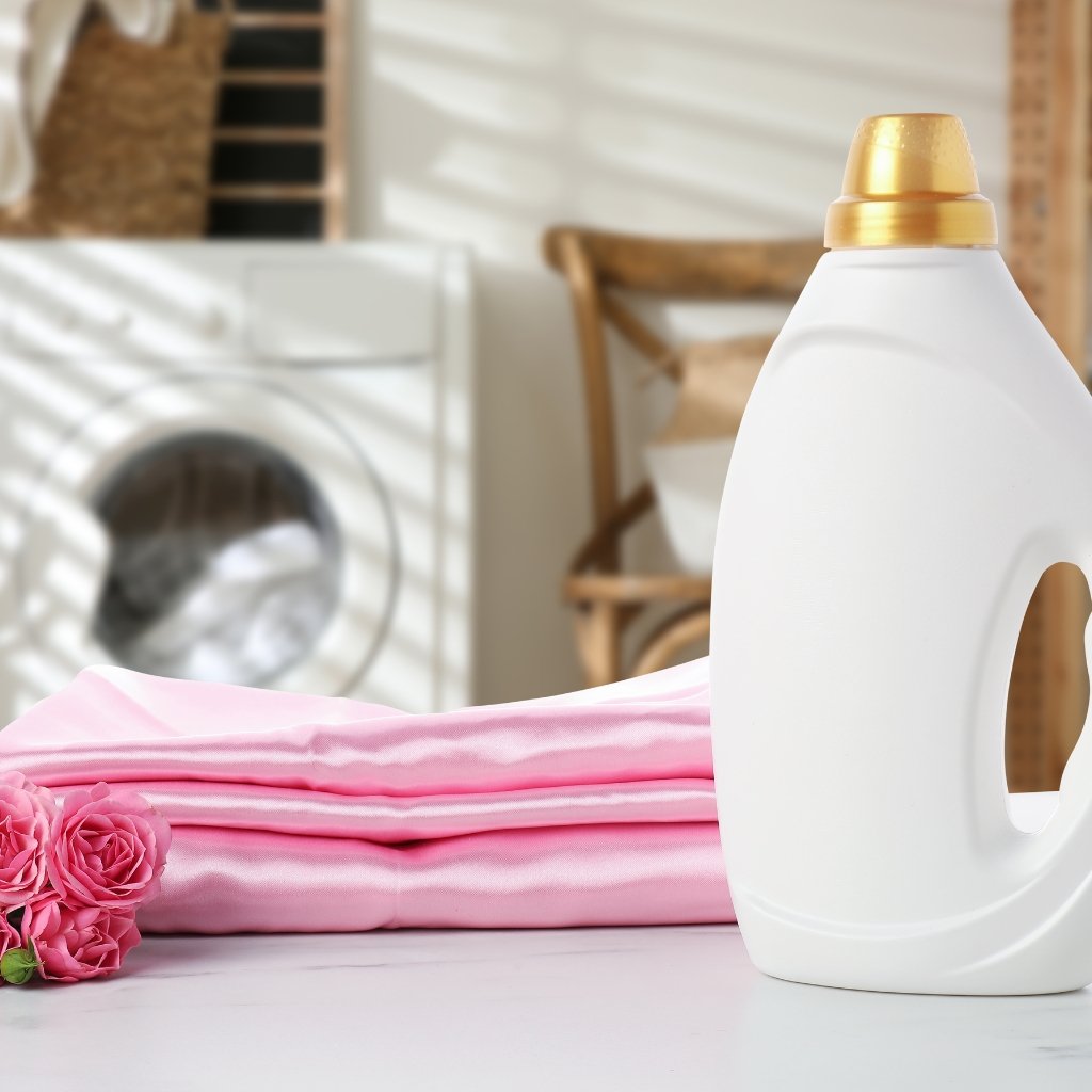 A white laundry detergent bottle with a gold cap sits on a surface beside folded pink clothes and pink flowers in a laundry room.