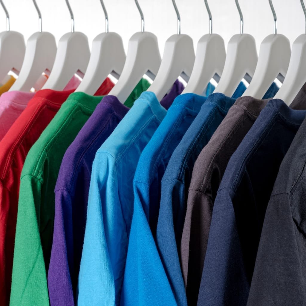 Row of colorful shirts on white hangers hanging in a retail display, arranged in a rainbow spectrum from red to dark blue.