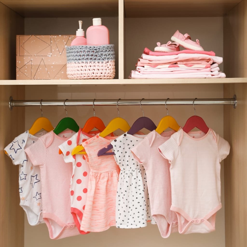 A wooden baby closet with a metal rod holding colorful baby onesies on bright hangers. On the top shelf there are pink bottles in a knitted basket and a stack of pink clothes, along with a folded box and baby shoes.