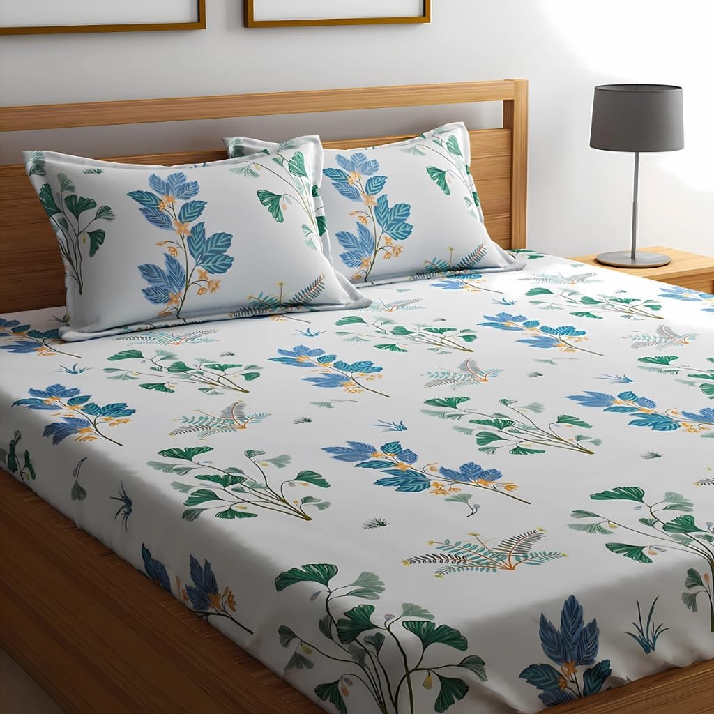 A neatly made bed with a wooden headboard and matching nightstand, covered by a white cotton bedsheet with a colorful botanical print in blue, green, and yellow. Two matching pillows rest against the headboard, and a modern gray table lamp sits on the nightstand.