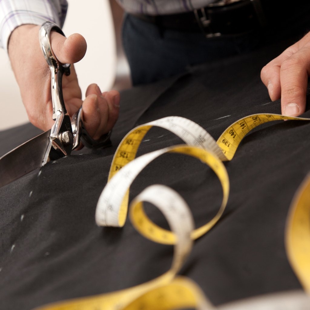 Close-up of a tailor’s hands cutting black fabric with metal shears, with a yellow measuring tape draped across the fabric.
