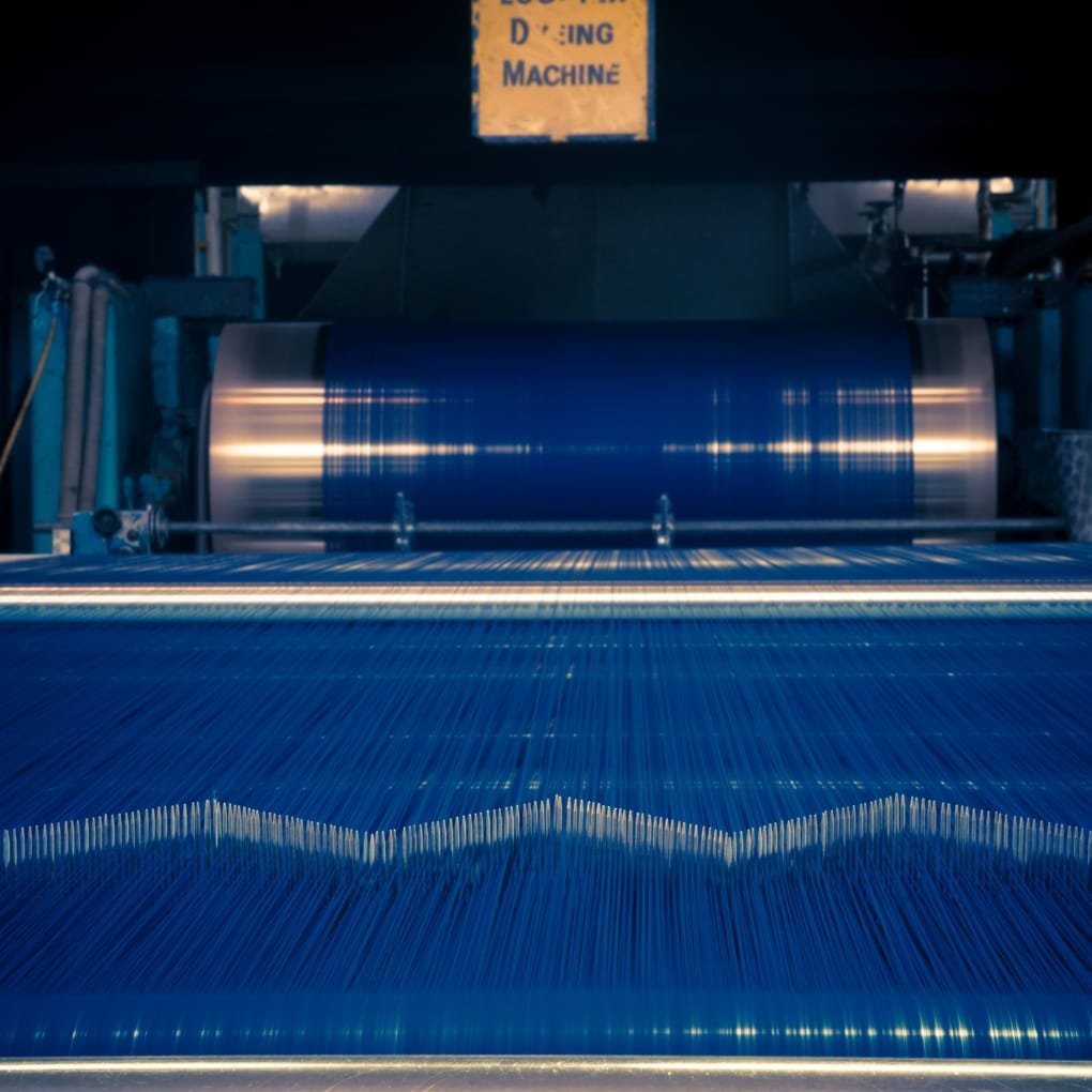 Industrial denim dyeing machine in operation with a large blue dye drum and parallel yarns creating a wavy pattern, dimly lit factory setting.