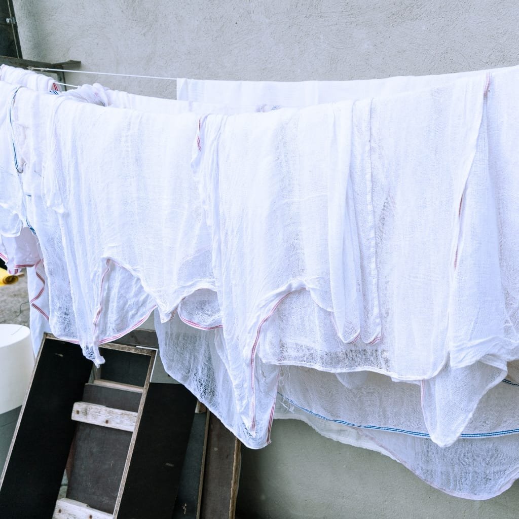 White household linens hanging on a clothesline to dry, including towels or sheets, with a wooden step ladder and a gray wall in the background. canvas cotton fabric