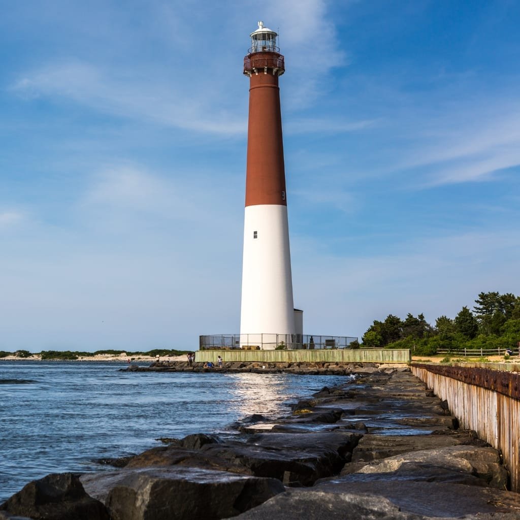 A tall, two-tone lighthouse with a brown upper section and white lower section stands on a rocky shoreline, with a calm sea and blue sky in the background.