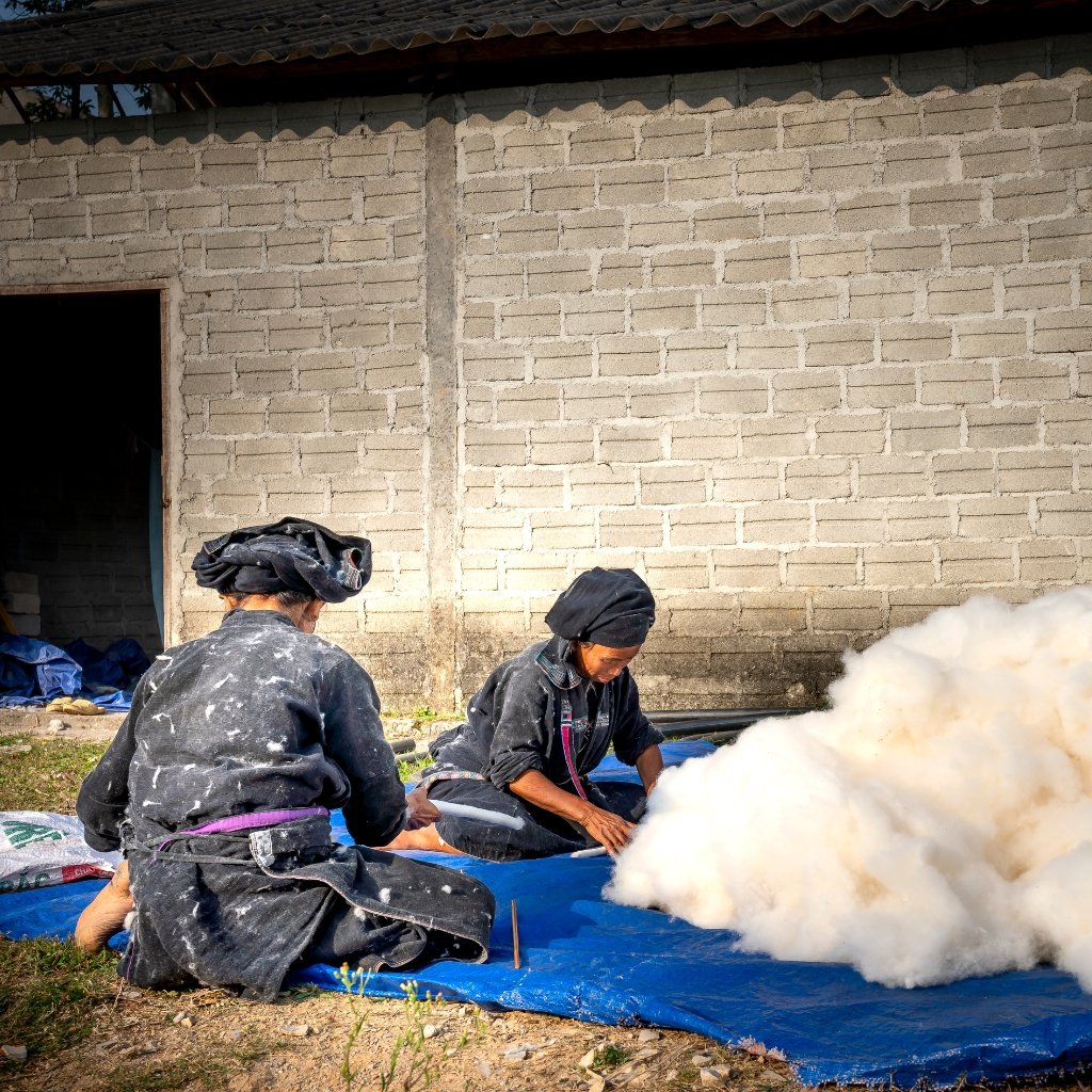 Two people wearing dark clothing and head coverings sit on a blue tarp outdoors next to a gray brick wall. One person appears to be handling white fluffy material, with a large cloud of dust or steam rising nearby.