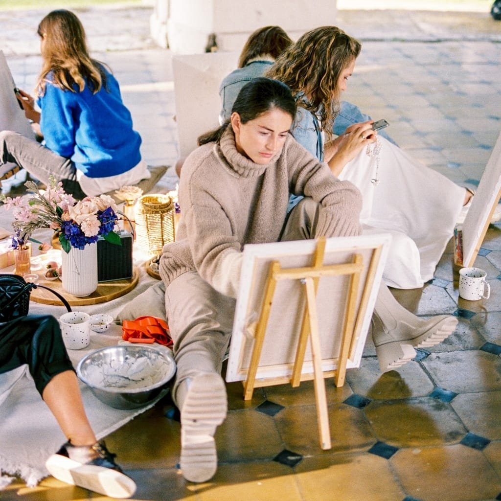 A group of young women drawing and chatting in a bright outdoor studio. One woman in a beige turtleneck sits on the floor, sketching at an easel, while others work on their projects nearby. Art supplies, a vase of flowers, and a cup are scattered around on the mosaic-tiled floor.