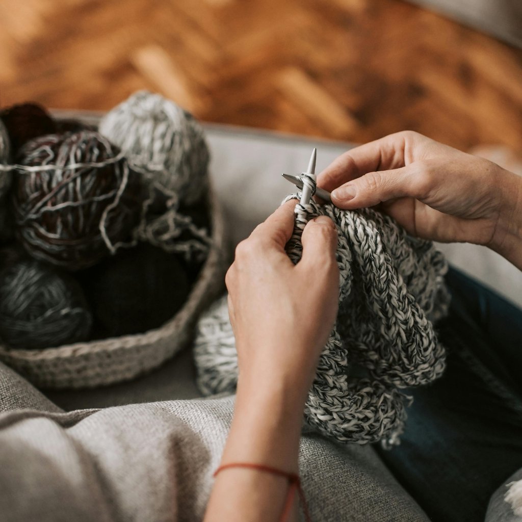 Close-up of hands knitting with thick gray yarn and metal knitting needles; a ball of yarn in a basket in the background.