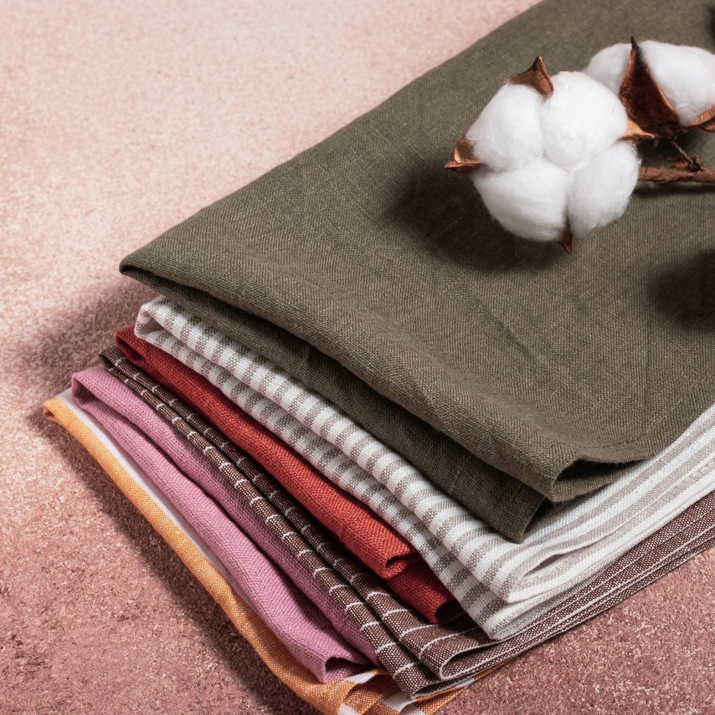 A stack of folded fabric in earthy tones (olive, taupe, beige, brown, burgundy, pink) arranged neatly on a pinkish surface with a white cotton boll resting on top.