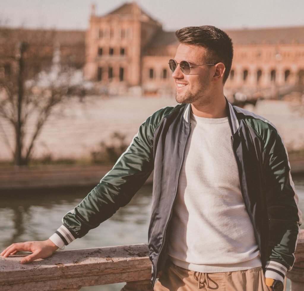 A stylish man stands outdoors near a stone railing by a canal, wearing a dark satin bomber jacket over a cream sweater, tan jogger-style pants, sunglasses, and a watch. He smiles and looks to the right with a cityscape in the softly blurred background. Best stain bomber jacket