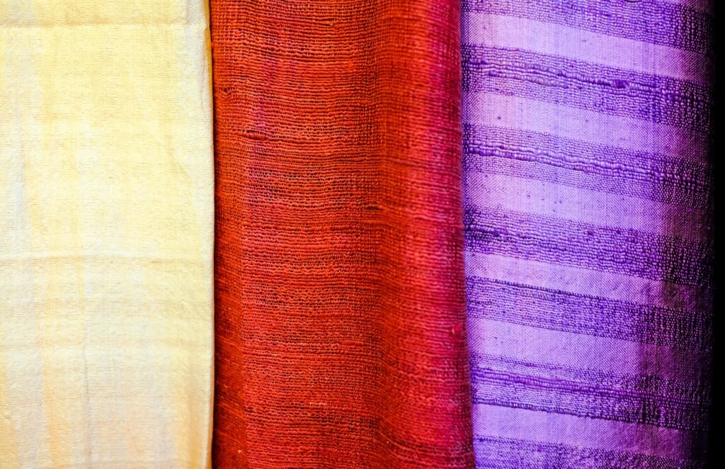 Close-up of three rolled fabrics in pale yellow, deep red, and purple with textured weaves.