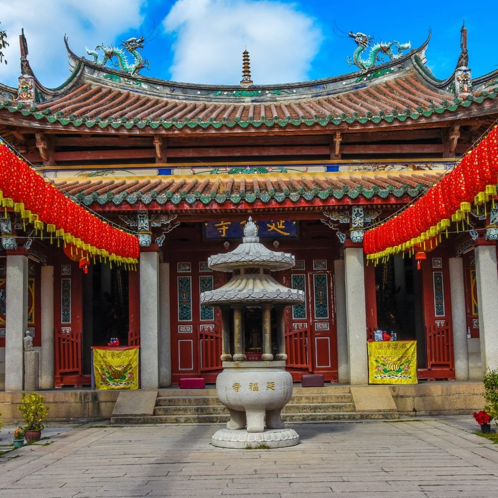 Temple courtyard with a stone incense burner centered in the foreground, red doors and decorative panels behind, festive red lantern garlands along both sides, and a traditional Chinese temple roof with dragon motifs against a bright blue sky.