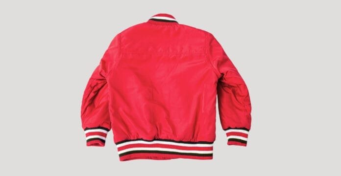 A bright red varsity bomber jacket viewed from the back, featuring ribbed cuffs and waistband with black, white, and red stripes. The jacket has gathered sleeves and a short collar with matching striped trim.