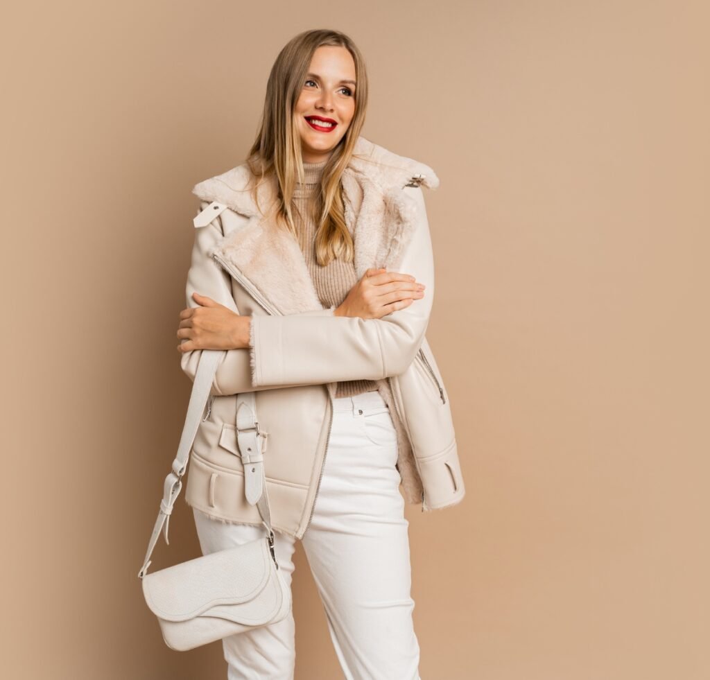 A stylish woman wearing a cream shearling bomber jacket, beige turtleneck, and white jeans, carrying a cream crossbody bag. She stands against a warm beige background with a bright, confident smile.