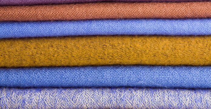 A stack of folded wool fabric in various colors (from top to bottom: reddish-brown, orange-brown, royal blue, mustard, medium blue, and light blue with a subtle pattern).