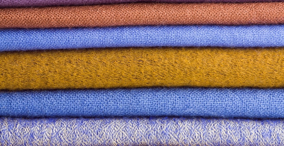 A stack of folded wool fabric in various colors (from top to bottom: reddish-brown, orange-brown, royal blue, mustard, medium blue, and light blue with a subtle pattern).