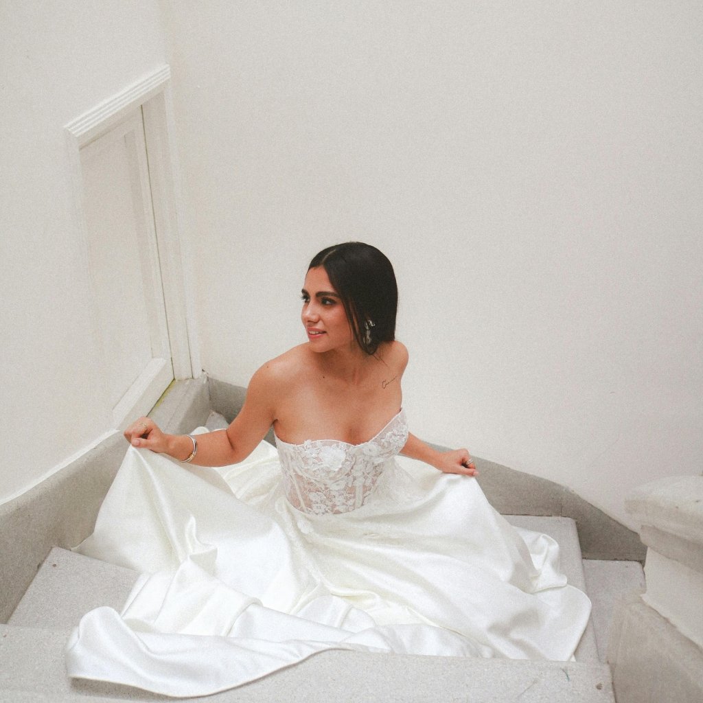 A woman sits on a white staircase wearing a strapless white satin-organza wedding gown with a delicate lace bodice. She gazes to the side, her dark hair pulled back, and holds the dress with both hands as it pools around her on the steps. Neutral white walls frame the scene.