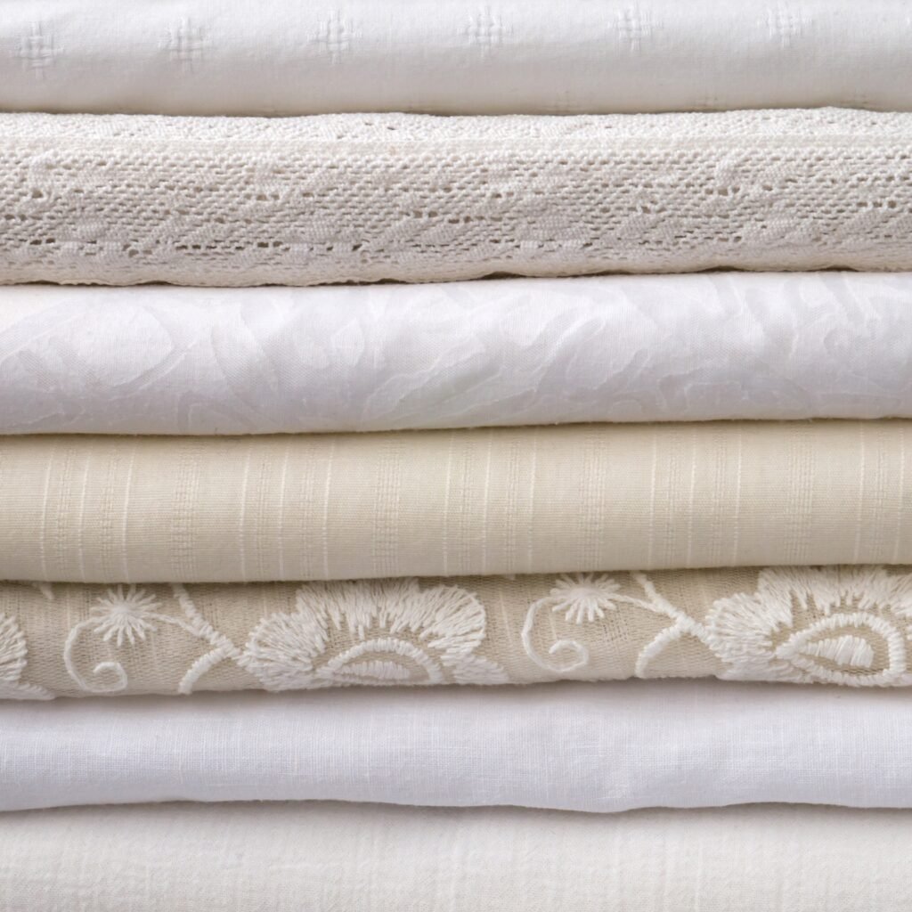 A stack of neatly folded lightweight fabrics in neutral tones (white, cream, ivory) with various textures including smooth plain weave, delicate lace, subtle stripes, and floral jacquard designs.