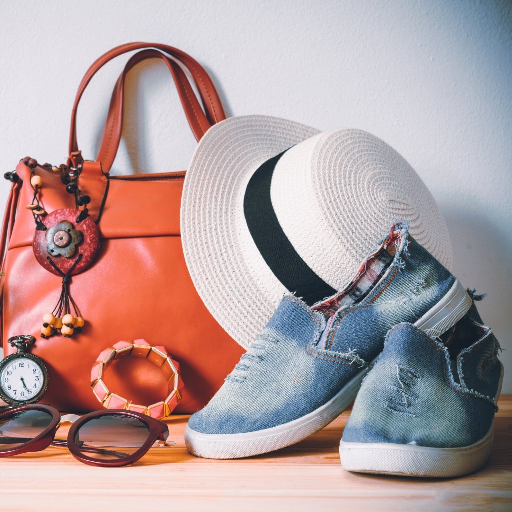 A stylish travel ensemble: a large orange handbag, a white straw hat with a black band, a pair of ripped denim high-top sneakers, a light blue plaid scarf, sunglasses, a pocket watch, and colorful jewelry on a wooden surface.