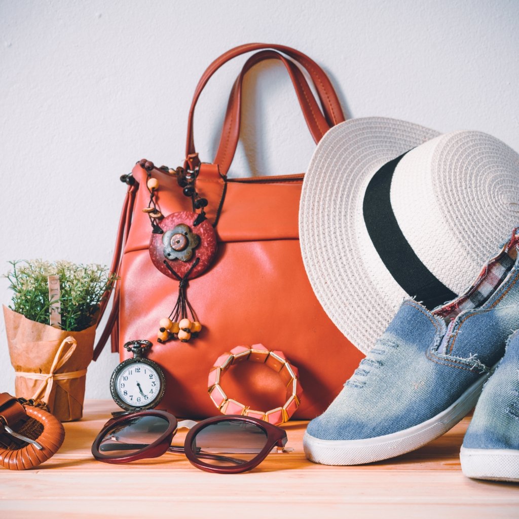 Orange handbag with decorative tassel and accessories, placed beside a straw hat, sunglasses, and jeans on a wooden surface.