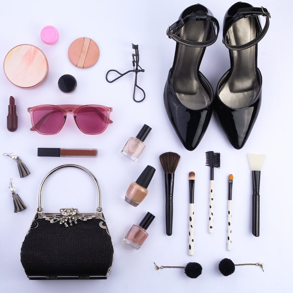 A flat lay of fashion accessories and beauty items on a light purple background, including black patent heels, pink sunglasses, lipstick, nail polishes, makeup brushes, a black beaded handbag, earrings, a compact powder, and various cosmetics arranged in a grid.