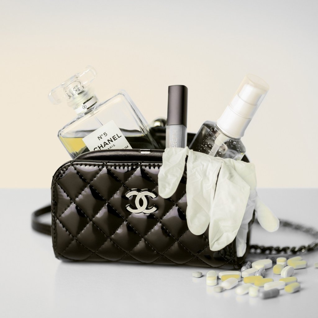 A black quilted Chanel handbag filled with beauty items and a pair of white gloves; various bottles (perfume, cosmetics) protrude from the open bag, with scattered white and yellow pills on the surface.