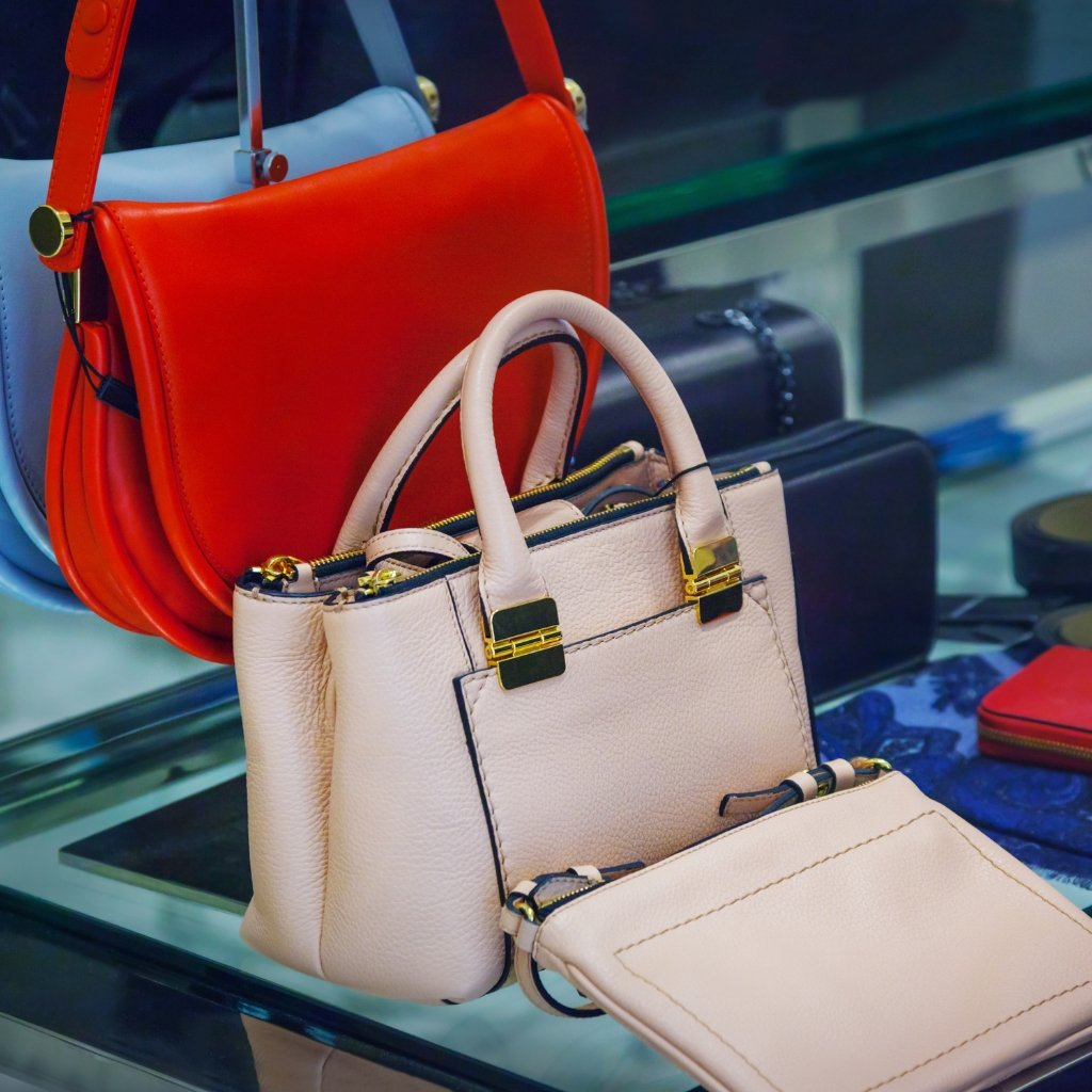 A display of fashion handbags, including a light beige tote with gold hardware and a matching small pouch, set against vibrant red and dark blue bags in a store window.