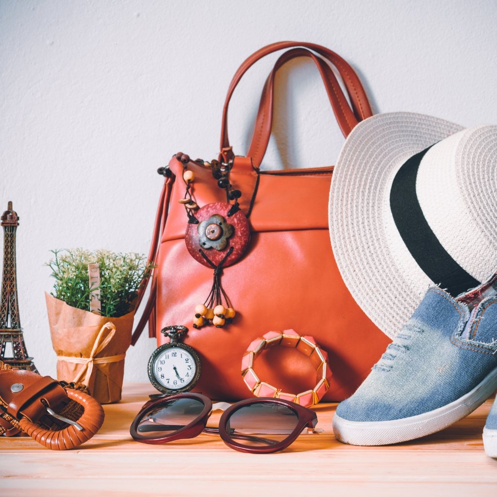 A flat lay of a stylish orange handbag with a decorative charm, a wide-brim hat, light blue sneakers, sunglasses, a small alarm clock, a bracelet, a belt, a potted plant, and a tiny Eiffel Tower figurine arranged on a wooden surface.