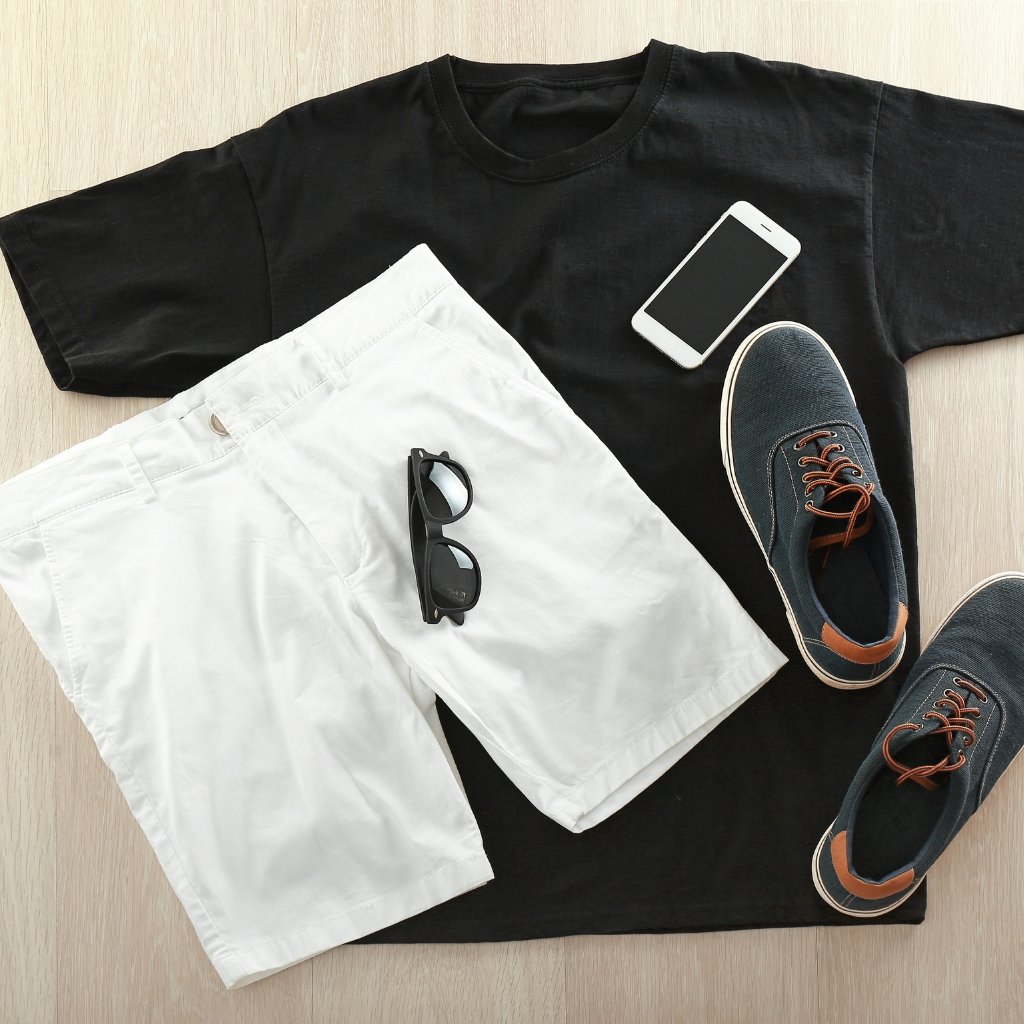 A flat lay outfit laid on a light wood floor, featuring a black short-sleeve T-shirt as the base, white shorts layered on top, a pair of dark blue sneakers with tan accents, black sunglasses resting on the shorts, and a white smartphone placed on the T-shirt.