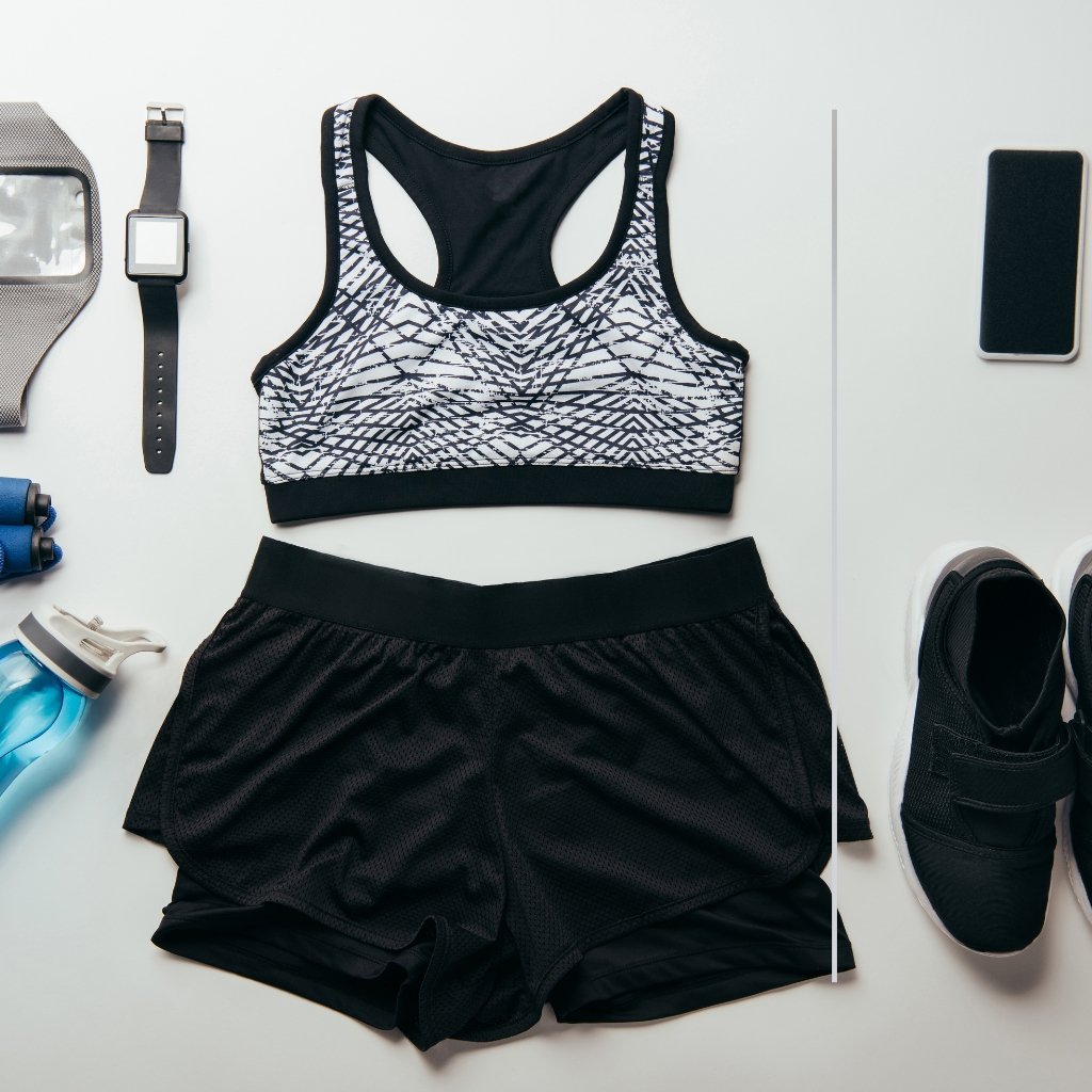 A flat lay of a black-and-white patterned sports bra, black athletic shorts, and surrounding gear including a smartwatch, water bottle, sneakers, and a smartphone.