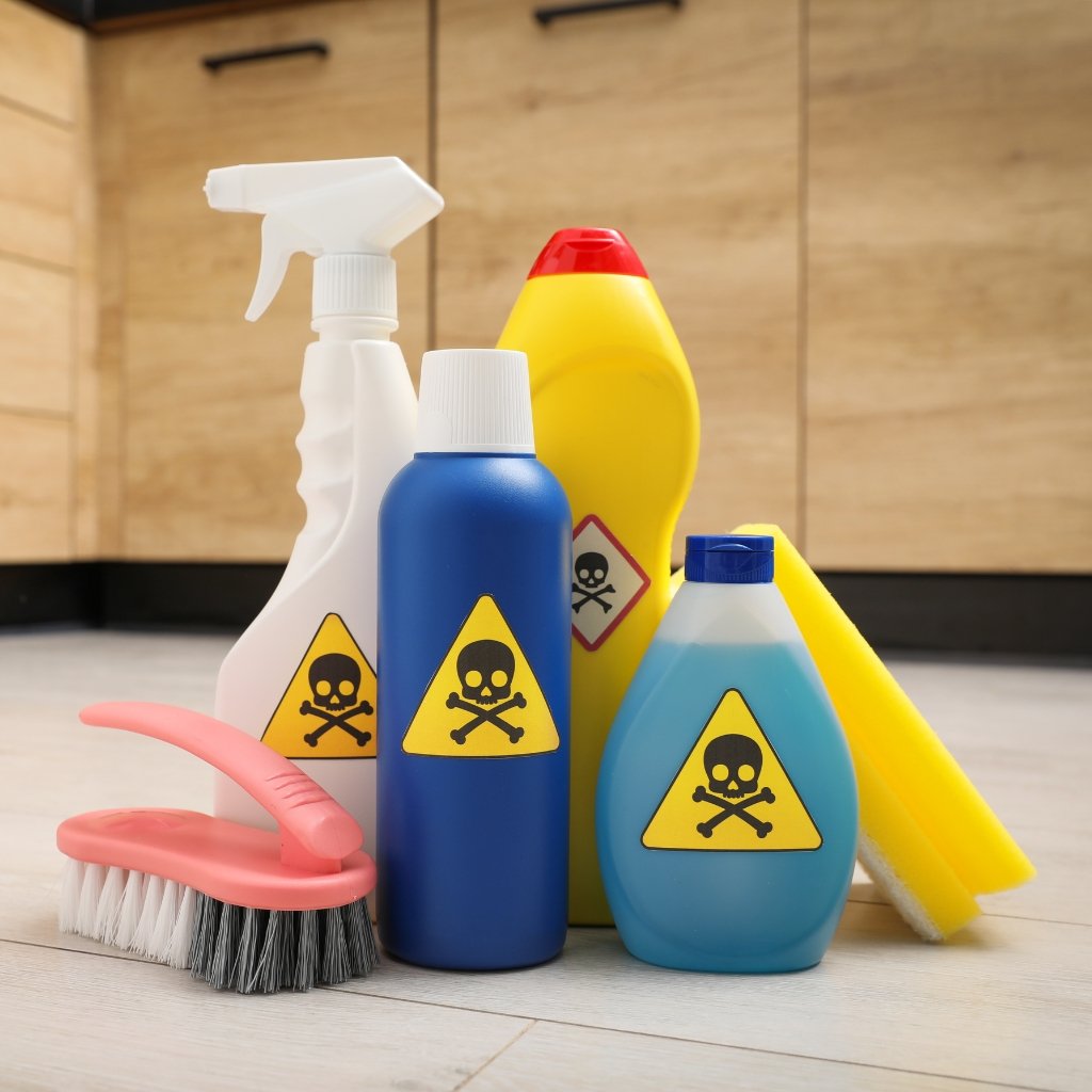 group of cleaning supplies on the floor, including spray bottles, a yellow bottle, a blue bottle, a pink scrubbing brush, and a blue bottle with hazard symbols featuring skull and crossbones.