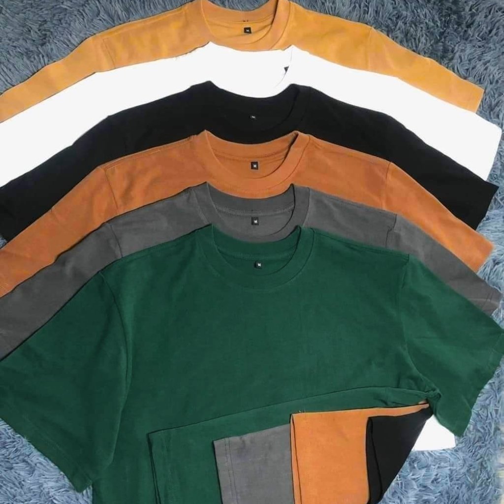 A top-down display of six oversized crew neck T-shirts in different colors—mustard yellow, white, black, burnt orange, gray, and dark green—layered in a fan shape on a blue fluffy surface. The bottom edges show small color swatches (orange, gray, and black).