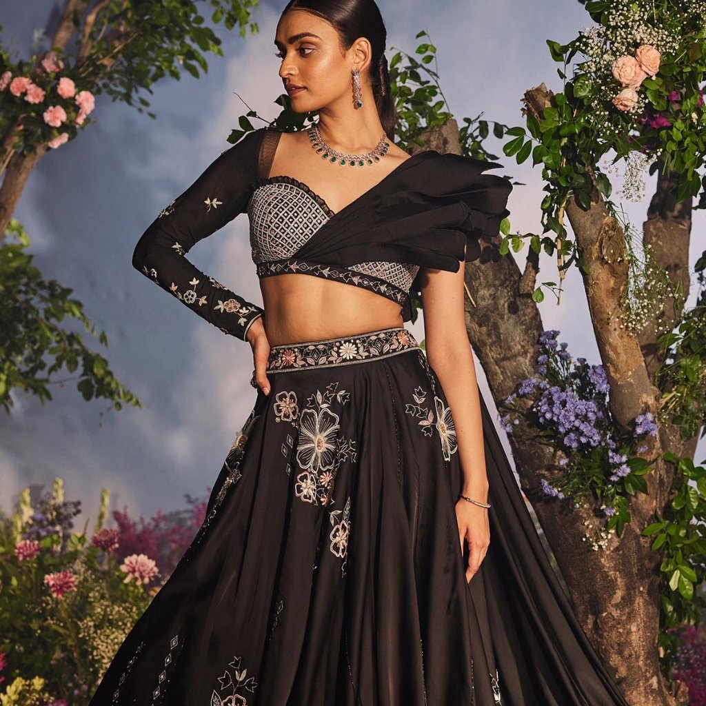 Black Organza Fabric: A Timeless Choice for Stylish Outfits 18 A woman wearing a black embroidered lehenga with a sheer, off-shoulder blouse and a matching dupatta, posing in a floral garden backdrop.