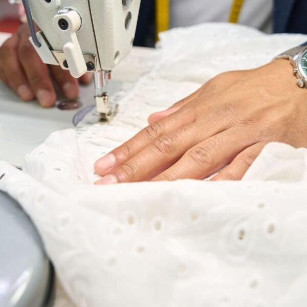 Close-up of a sewing machine stitching white fabric with a person's hand guiding the material; a wristwatch is visible on the hand.