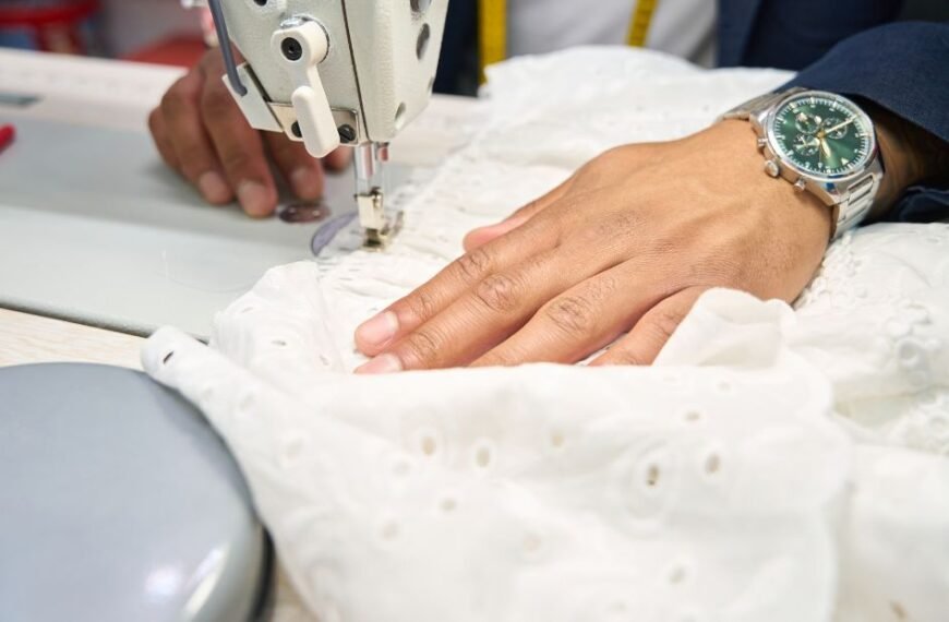 Close-up of a sewing machine stitching white fabric with a person's hand guiding the material; a wristwatch is visible on the hand.
