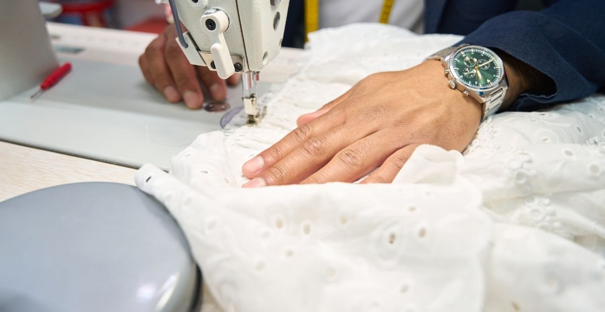 Close-up of a sewing machine stitching white fabric with a person's hand guiding the material; a wristwatch is visible on the hand.