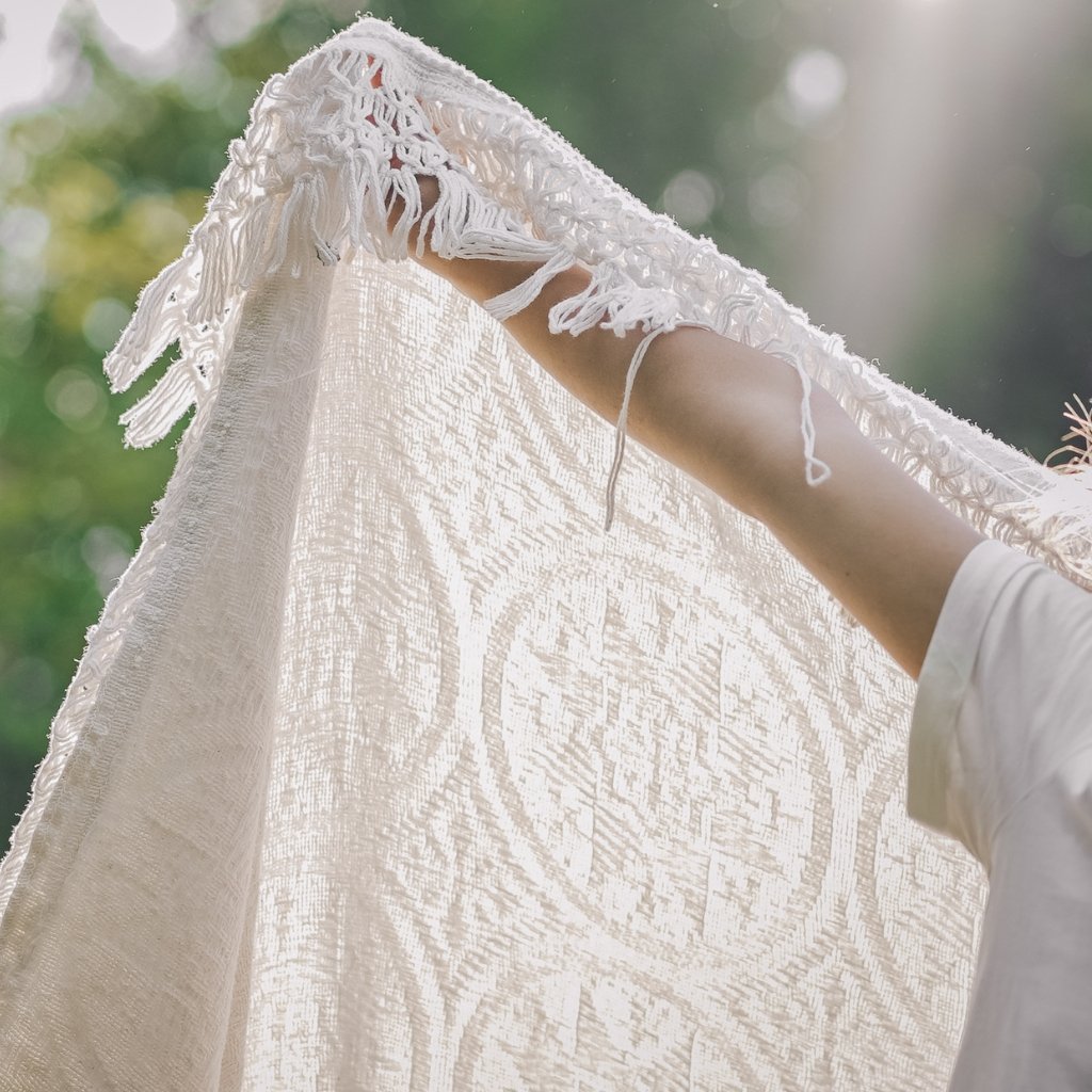 A close-up of a person holding up a delicate white lace or woven textile outdoors, with sunlight filtering through and a blurred green, natural background.