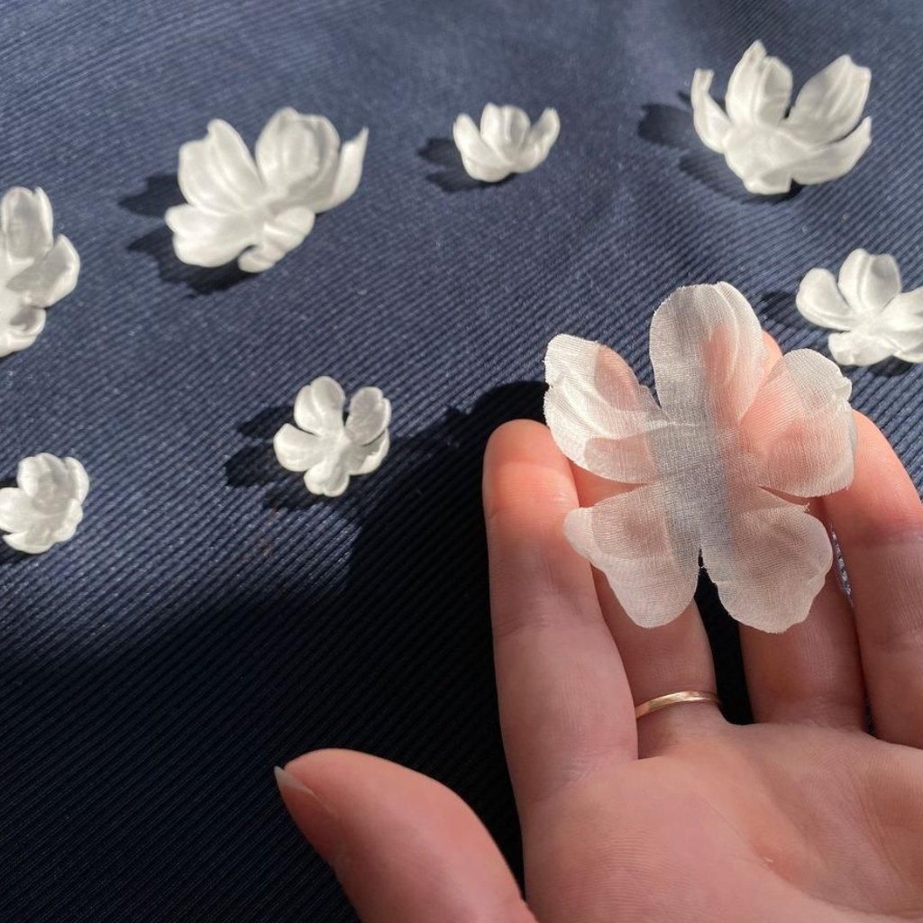 Close-up of a delicate, semi-translucent white silk organza flower being held between a person’s fingers. Several other small white fabric flowers sit scattered on a dark blue textured surface in the background