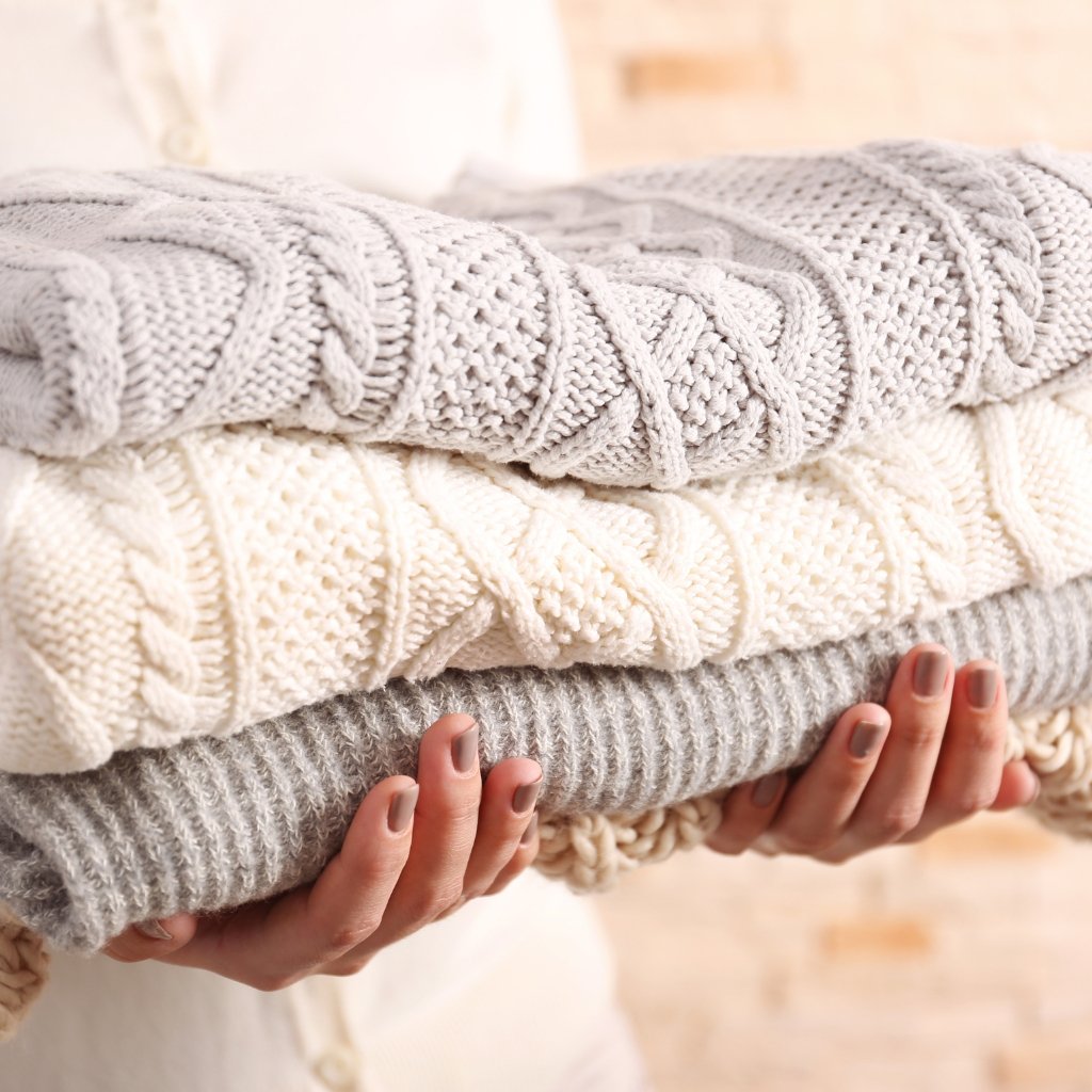 A person is holding a stack of thick, knitted blankets in neutral tones (gray and cream). The blankets show chunky cable and textured knit patterns, and the person’s hands with light gray nail polish support the bottom blanket.