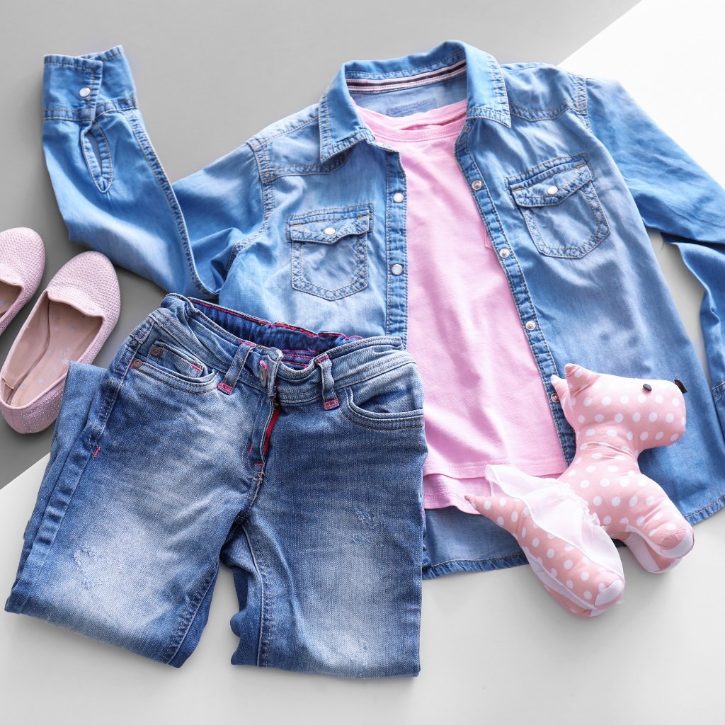 A flat lay of a denim outfit laid out on a light gray surface, including a light pink tee under a blue denim shirt, matching blue denim jeans, pink polka-dot stuffed animal, soft pink slip-on shoes, and no visible wool fabric.