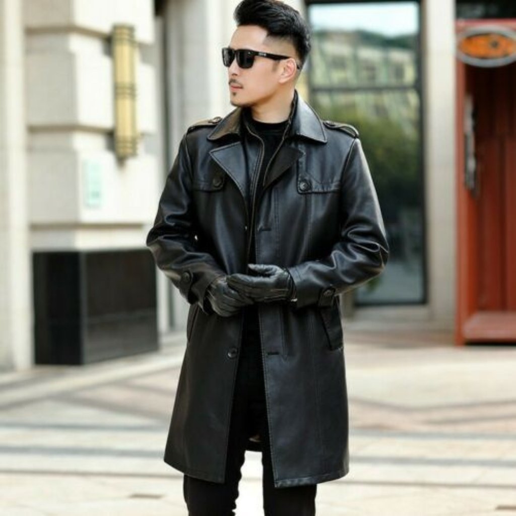 A stylish man stands outdoors wearing a long black leather duster coat, black gloves, and dark sunglasses, posing with his hands clasped. He has a neatly groomed beard and hair, urban backdrop with buildings in the background.
