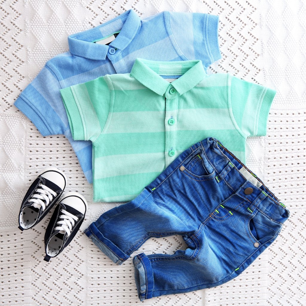 Set of baby boy clothes laid out on a light woven surface. Two polo shirts in pastel stripes (one blue, one mint green) with buttons, a pair of small denim shorts, and a pair of black-and-white sneakers. The fabrics appear soft and plush with a terry-wool-like texture in the clothing pieces.