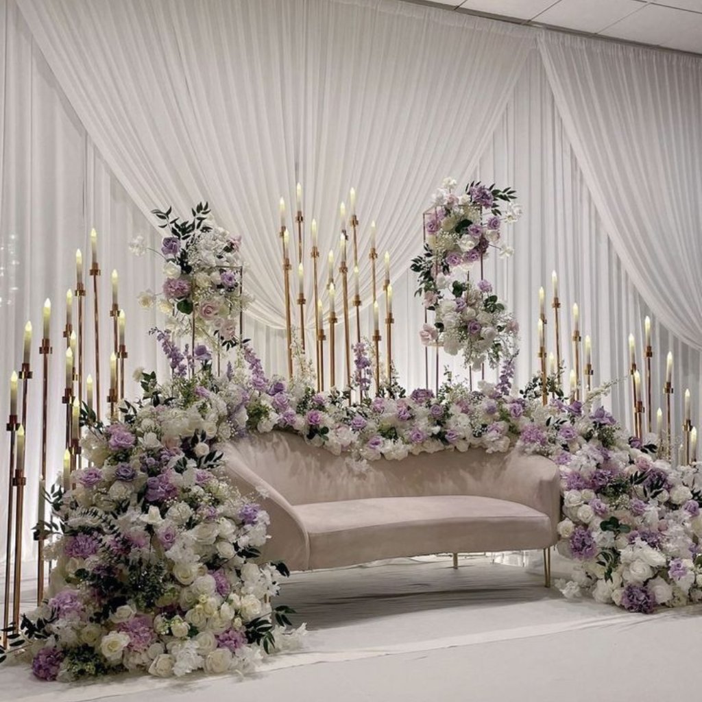 An elegant wedding stage with a blush sofa surrounded by lush white and pastel purple flowers, cascading floral arrangements, and tall gold candlesticks set against white draped backdrop.
