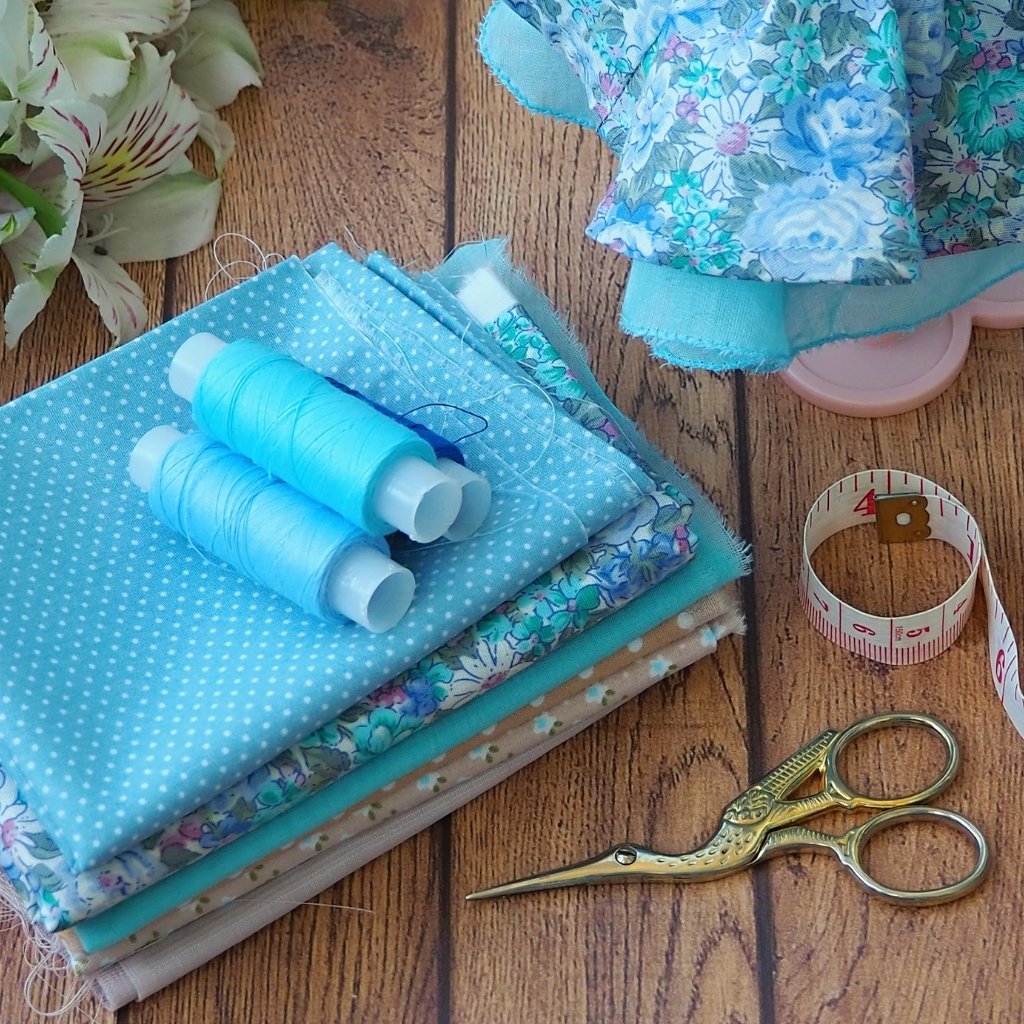 Dimity Cotton Fabric: Soft, Durable, and Timeless Choice 6 Sewing workspace with folded blue fabrics, spools of blue thread, measuring tape, decorative scissors, and fabric pieces on a wooden surface. The image shows a line of pastel-toned dresses on wooden hangers, suspended from a white metal rack. The warm sunlight creates delicate shadows and highlights the fabric textures, giving a calm, airy store or dressing-room vibe. A plant with long, slender leaves is visible in the foreground on the right, adding a natural touch.